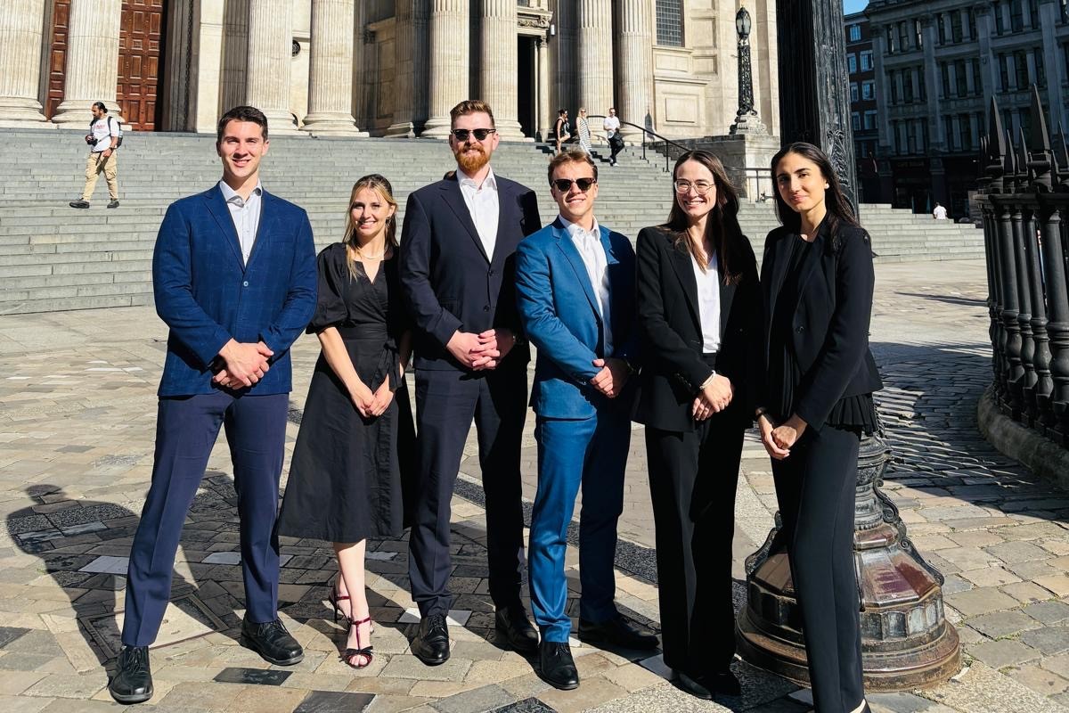 Six male and female law students in business attire, posing for a group photo in front of a tall, ornate building with pillars